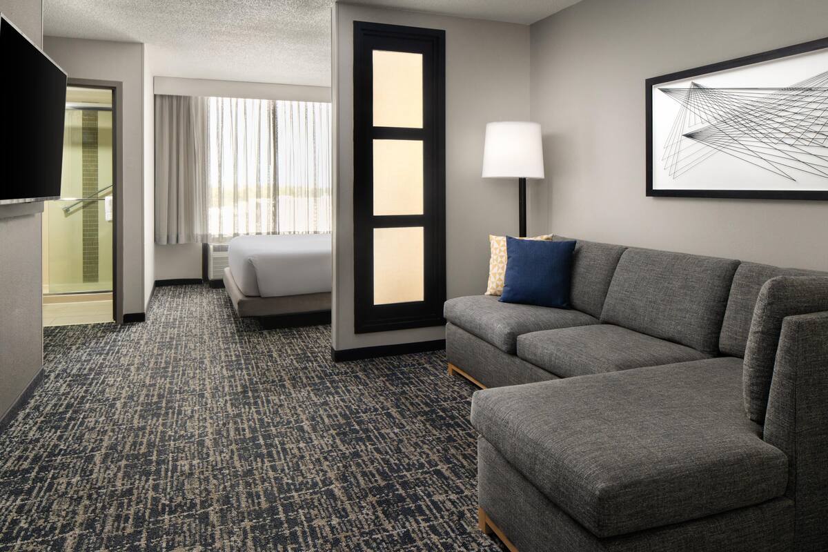 Ein Hotelzimmer mit grauem Sofa, einem Bett, einem Fernseher, einem Fenster mit Vorhängen, einer Glastür und einem gerahmten abstrakten Kunstwerk an der Wand.