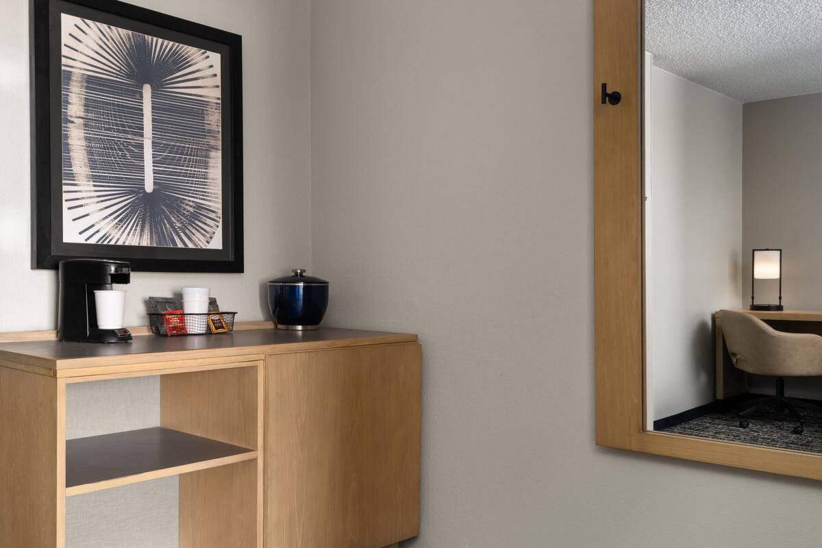 Ein Hotelzimmer mit Schreibtisch aus Holz, einem gerahmten Kunstwerk, einer Kaffeemaschine und einem Spiegel, der einen Stuhl und eine Lampe reflektiert.