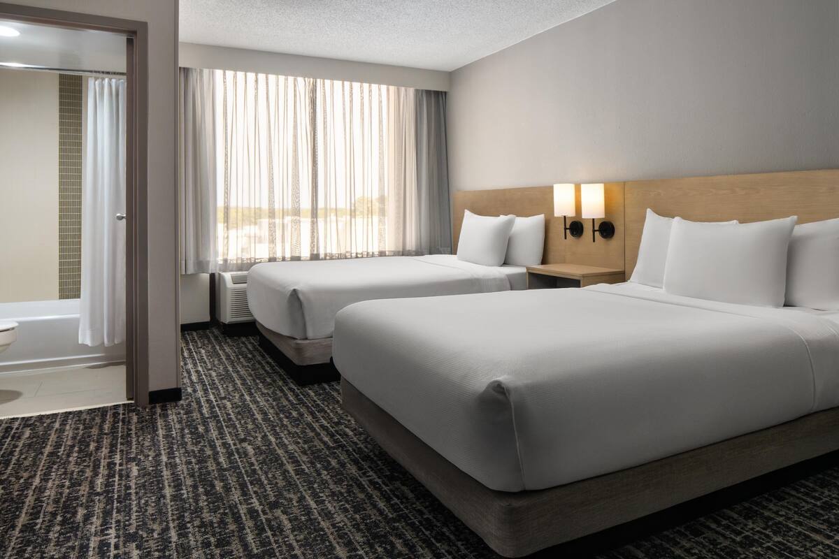 Ein Hotelzimmer mit zwei Betten, einem Badezimmer und einem großen Fenster mit Vorhängen.