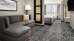 Habitación de hotel con sofá gris, cama, televisión y un ventanal con cortinas.