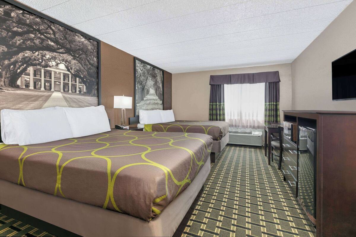 A hotel room with two beds, a desk, a chair, a TV, and a large window with curtains.