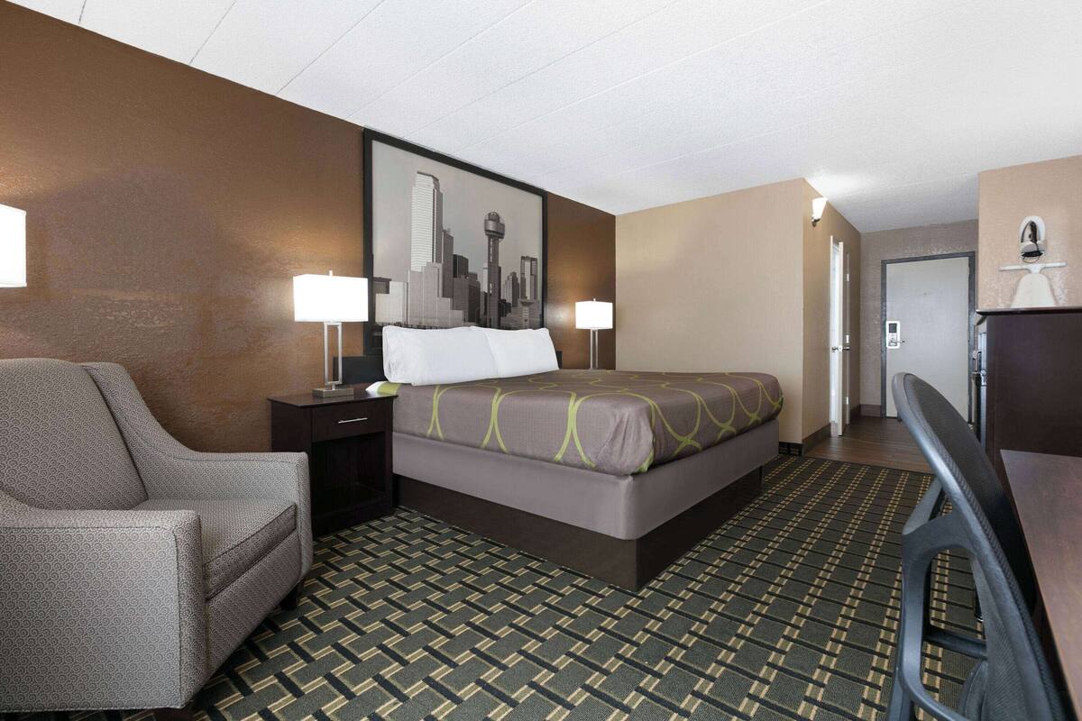 A hotel room with a bed, a chair, a desk, and a cityscape painting on the wall.