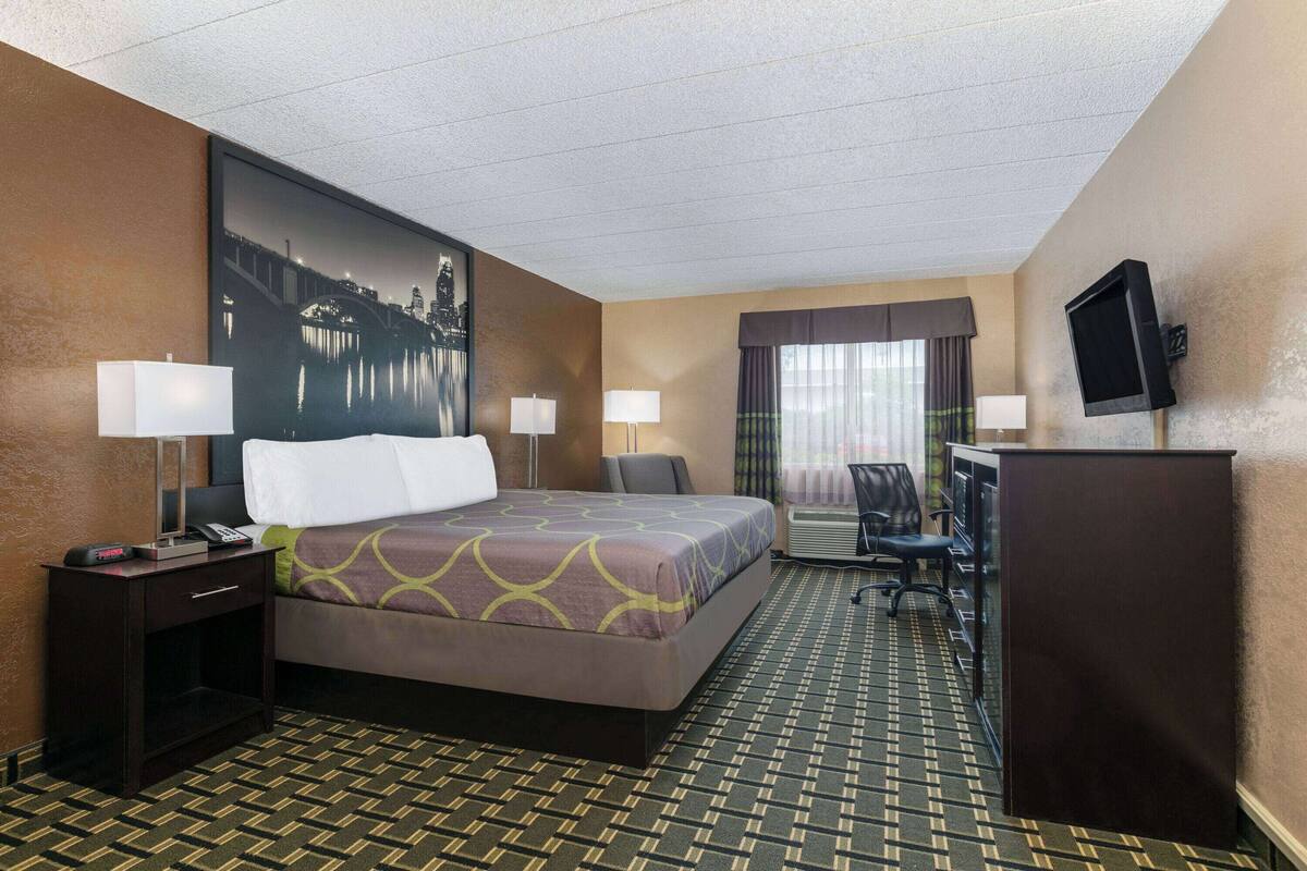 A hotel room with a bed, two bedside tables with lamps, a desk with a chair, a television, and a large window with curtains.