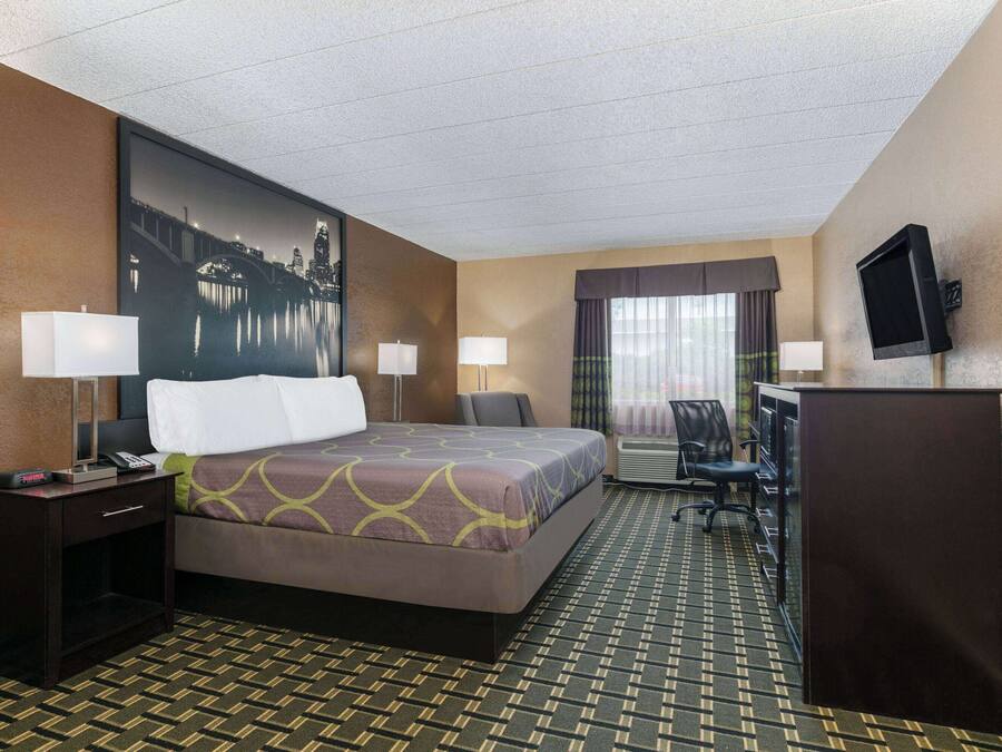 A hotel room with a bed, two bedside tables with lamps, a desk with a chair, a television, and a large window with curtains.