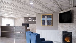 A hotel lobby with a reception desk, a blue sofa, a fireplace, and a TV mounted on the wall. - AmericInn by Wyndham Woodstock IL (Woodstock)