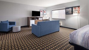 A hotel room with a patterned carpet, two sofas, a bed, a TV, a small table, and a window with blinds. - AmericInn by Wyndham Woodstock IL (Woodstock)