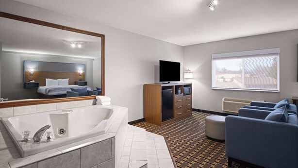 A modern hotel room with a large mirror, a bathtub, a flat-screen TV, a sofa, and a large window. - AmericInn by Wyndham Woodstock IL (Woodstock)