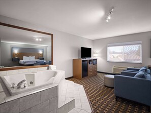 A modern hotel room with a large mirror, a bathtub, a flat-screen TV, a sofa, and a large window. - AmericInn by Wyndham Woodstock IL (Woodstock)