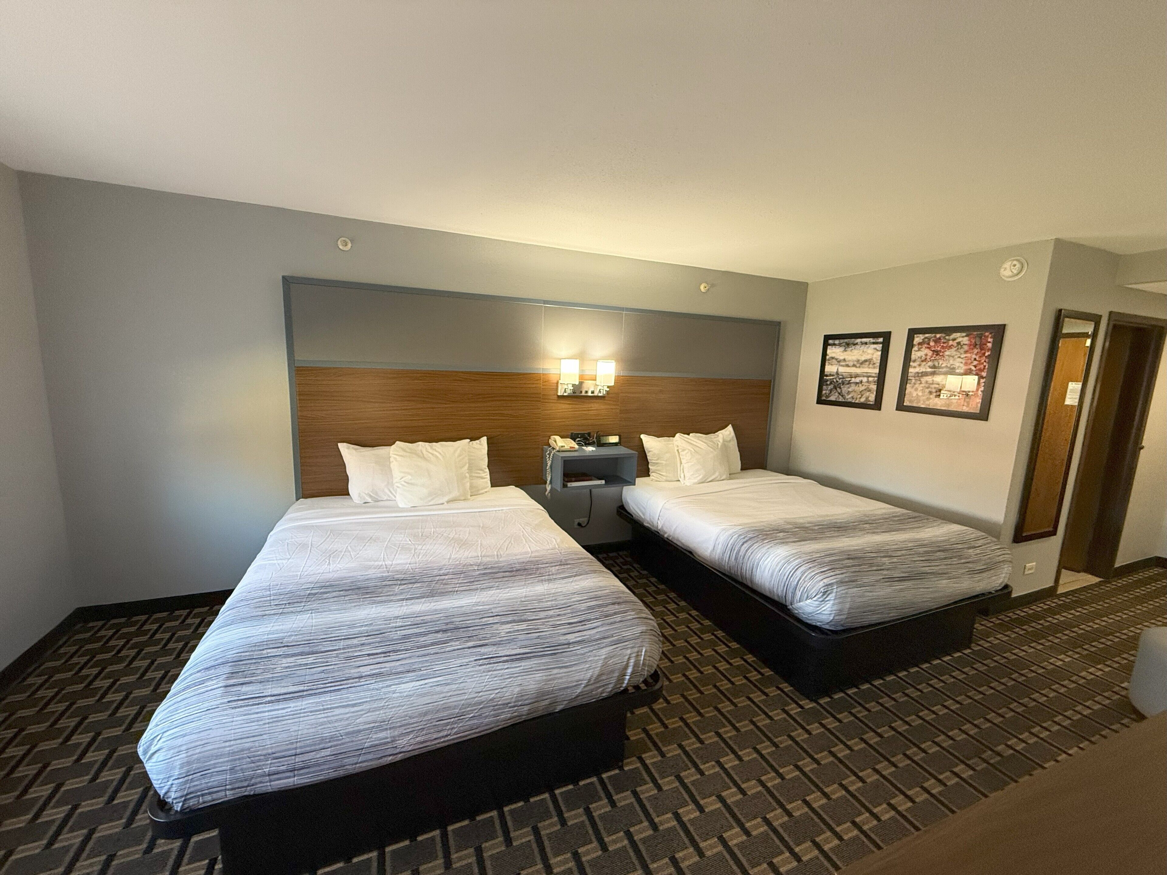 Habitación de hotel con dos camas, una mesita de noche con lámpara e iluminación en la pared.