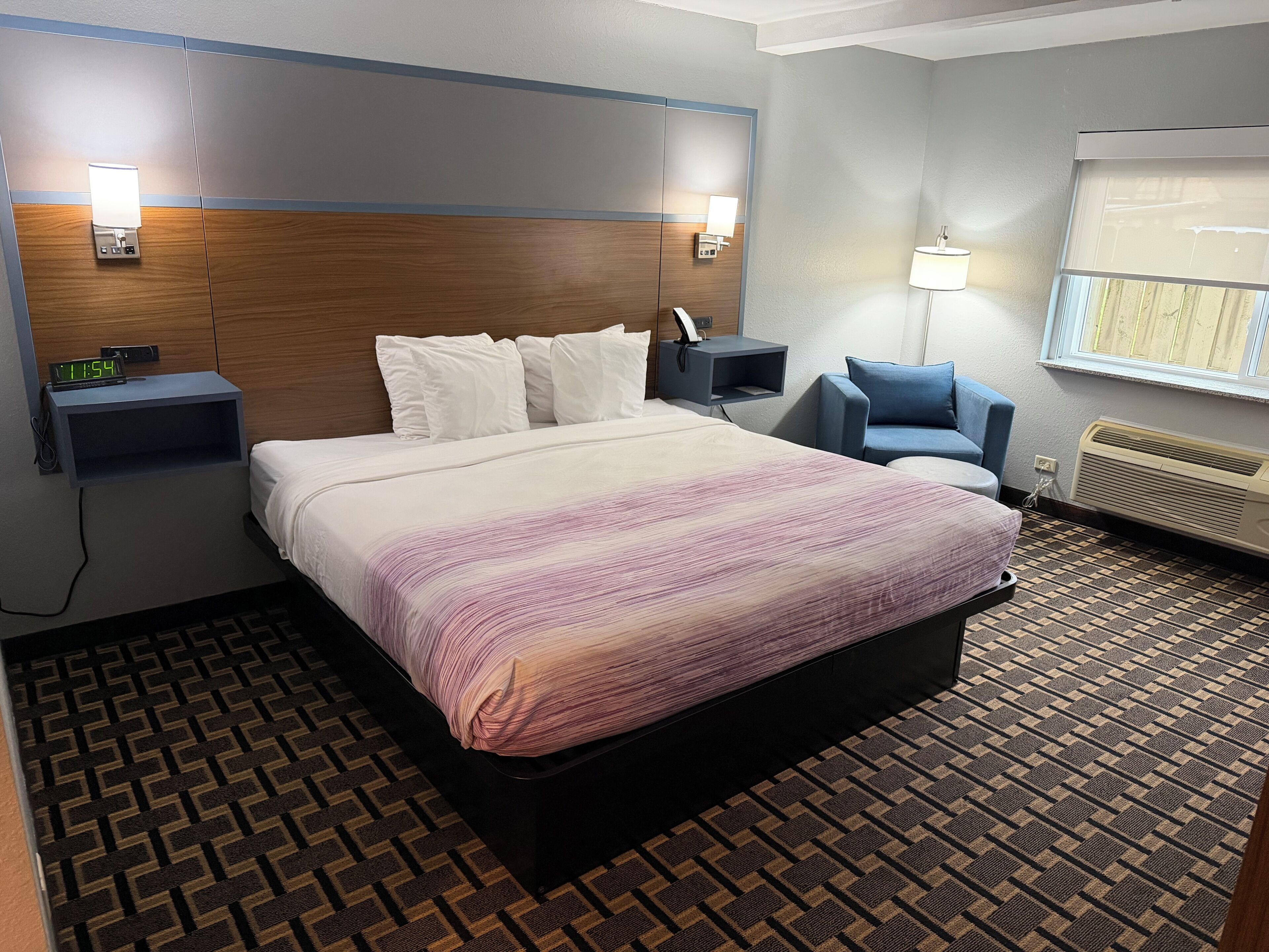 A hotel room with a bed, bedside tables, a chair, a window, and a wall-mounted heater.