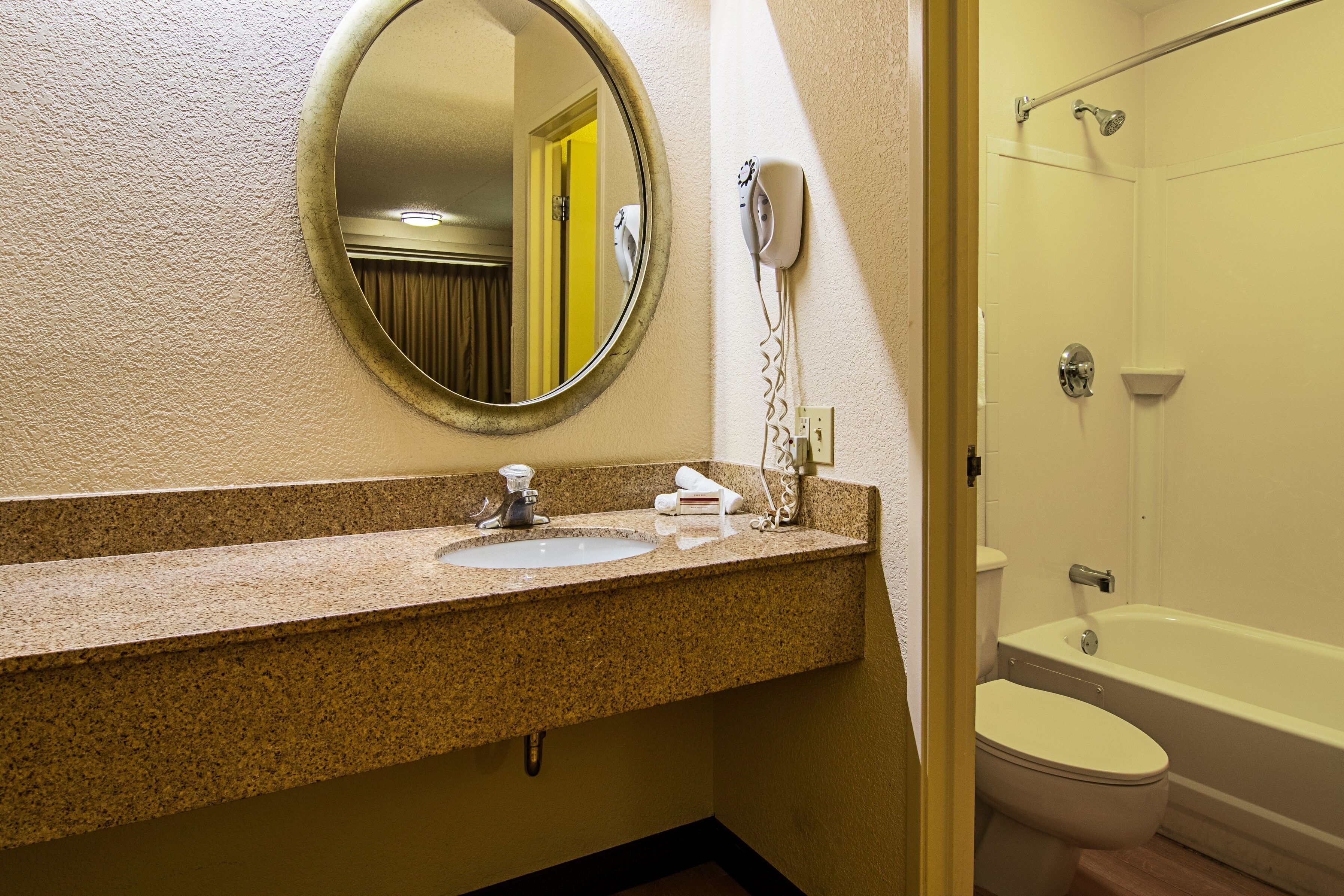 A bathroom with a granite countertop, a round mirror, a hairdryer, a sink, and a bathtub.