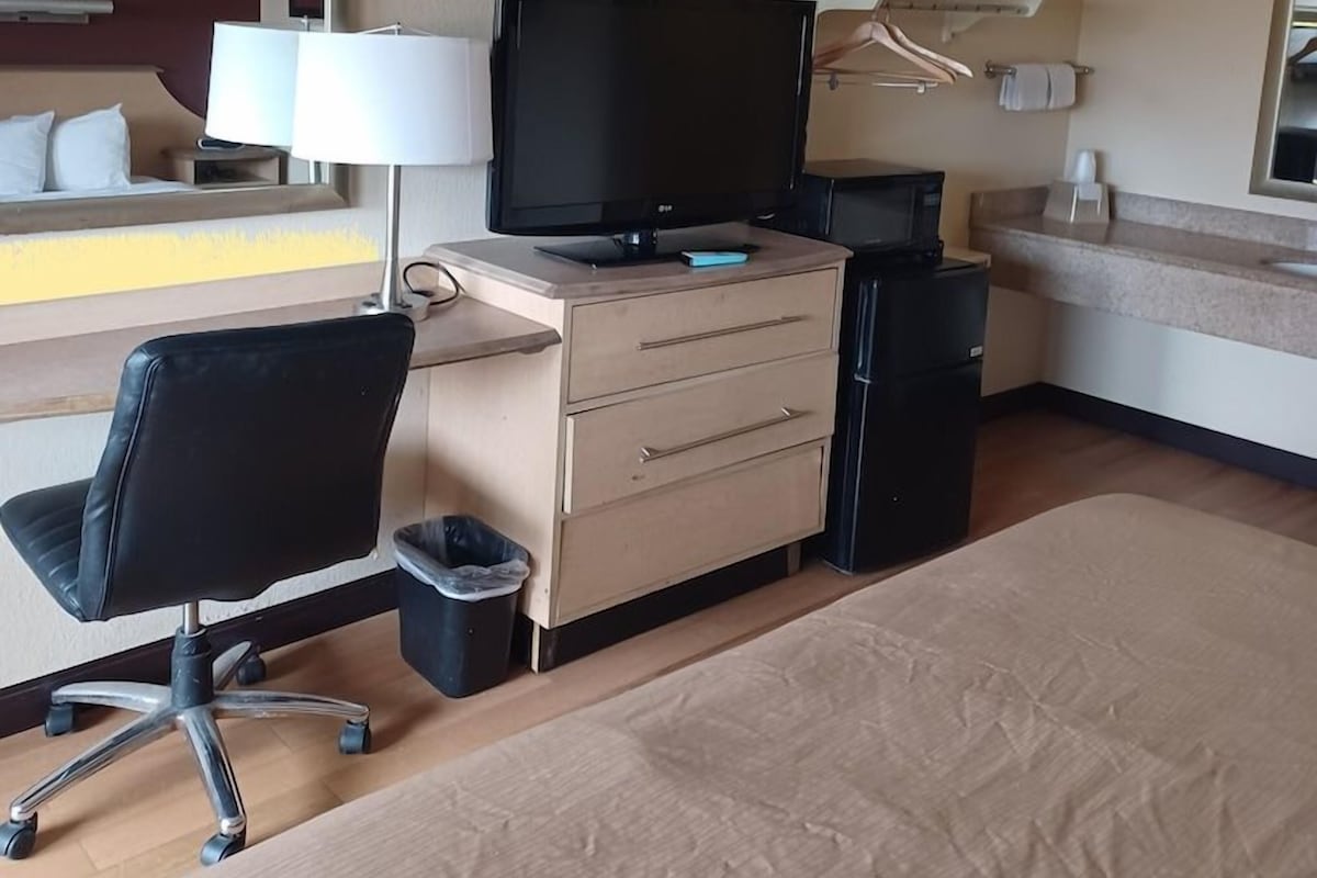 A hotel room with a bed, a desk with a television, a chair, a small trash bin, and a mirror.