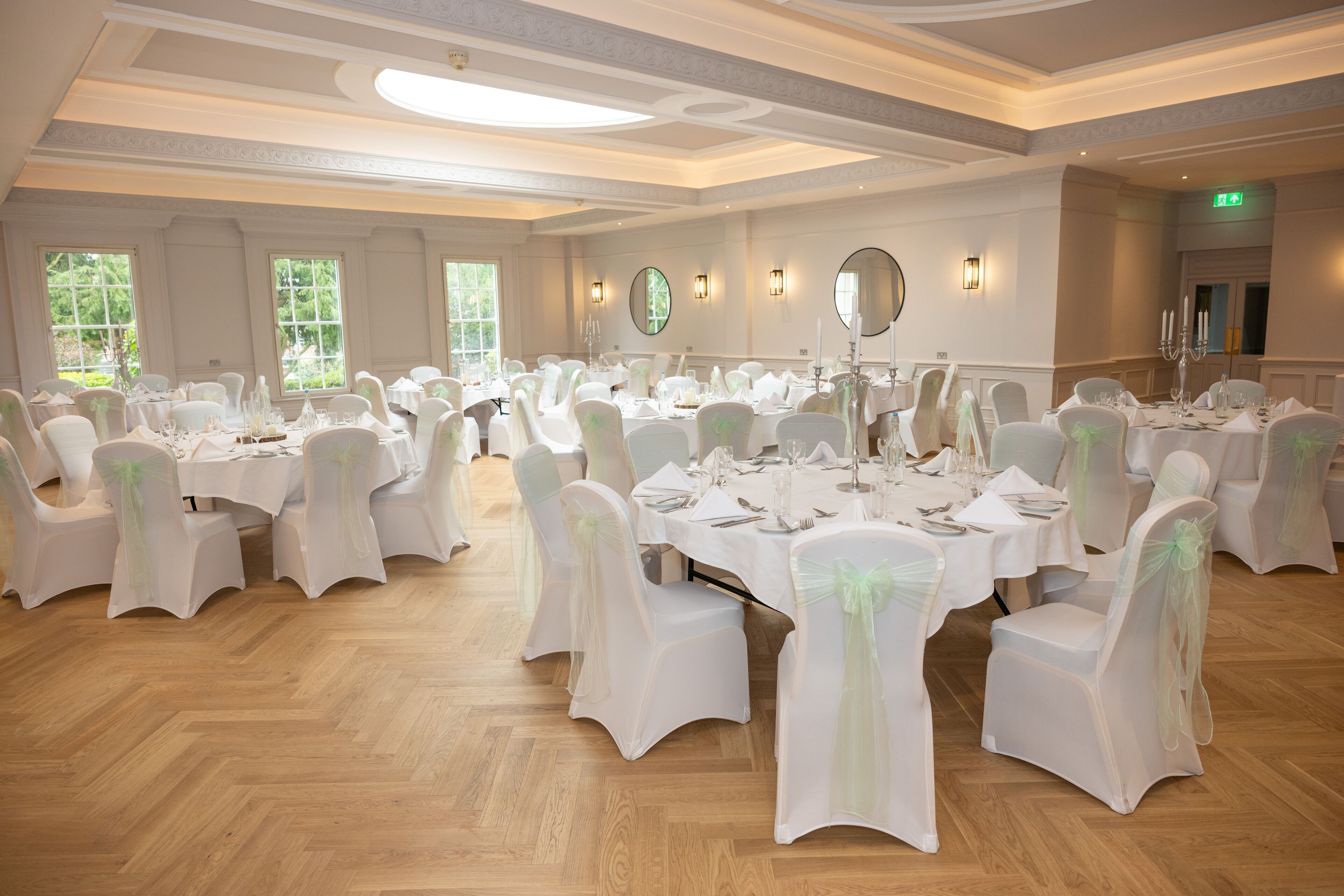 Un amplio salón de banquetes con varias mesas redondas dispuestas para comer, sillas blancas con cintas verdes y candelabros.