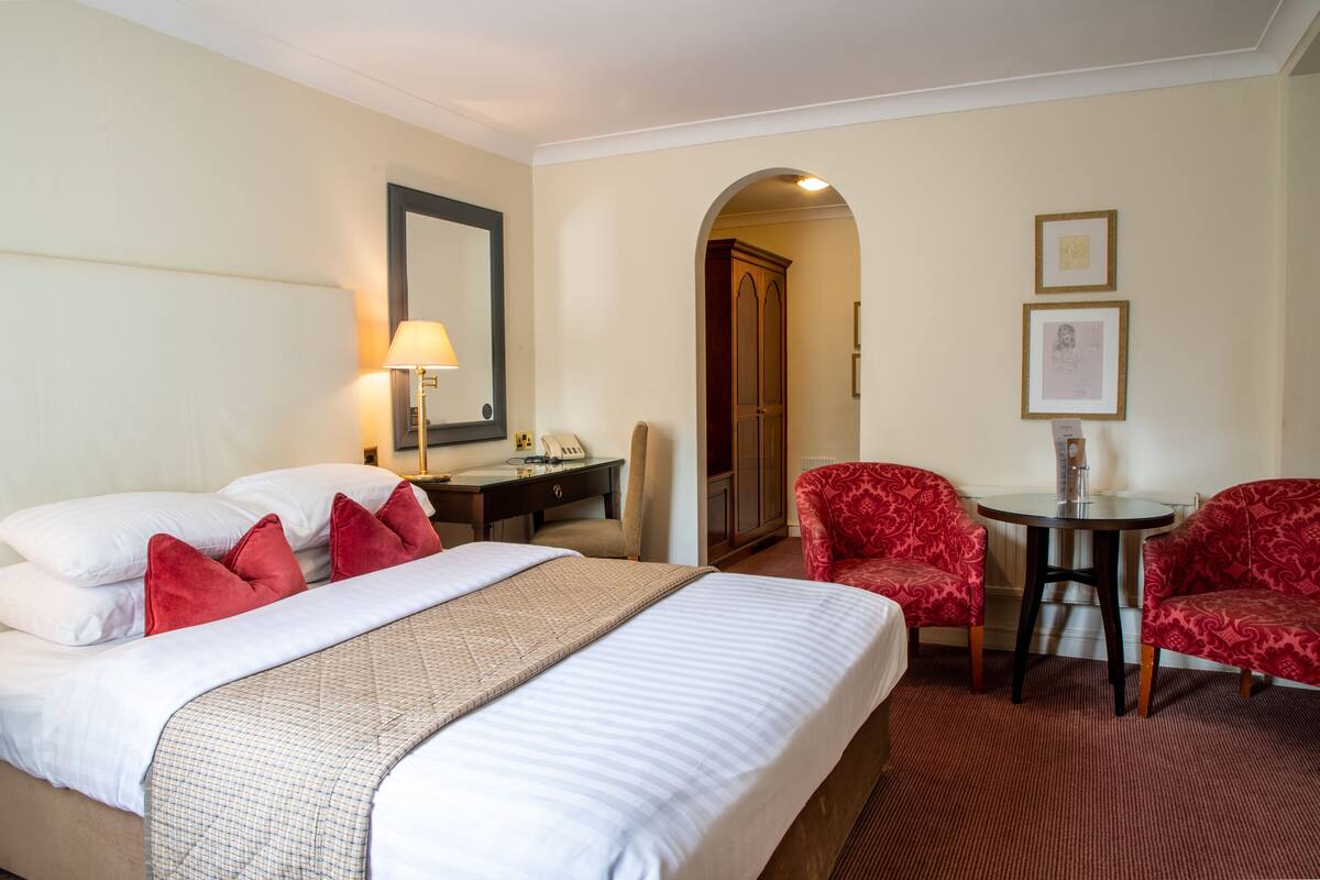 A hotel room with a large bed, a desk, two red armchairs, a small table, and a mirror.