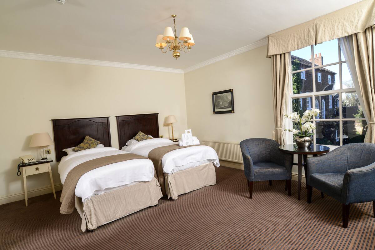 A hotel room with two beds, a sitting area with a table and chairs, a chandelier, and a window with curtains.