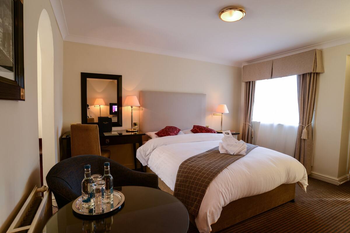 A hotel room with a bed, bedside tables, lamps, a chair, and a table with bottles.
