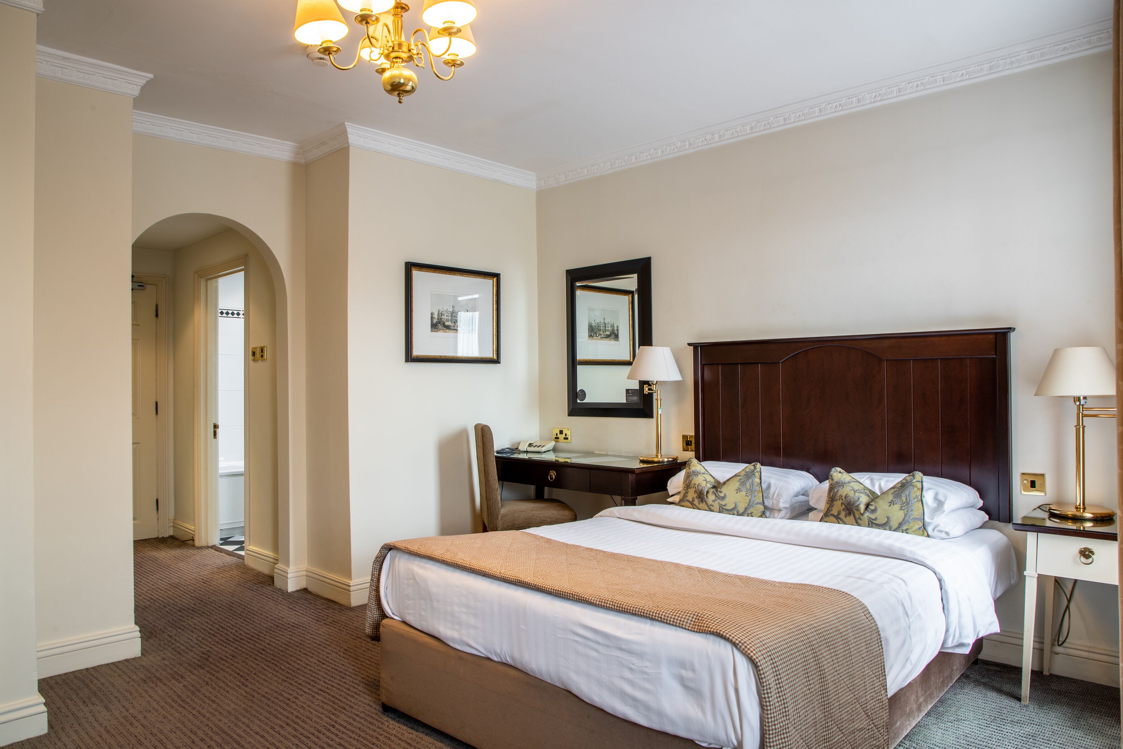 A hotel room with a large bed, a desk, a chair, a desk lamp, a bedside table, and a mirror.