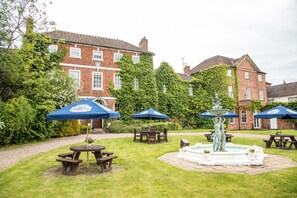 A courtyard with picnic tables and umbrellas, a fountain, and a brick building with ivy. - Park House Hotel (Shifnal)