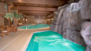 Piscina interna com cachoeira, bancos de madeira e parede de tijolos.