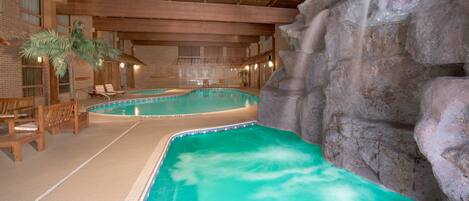 Una piscina interior con una cascada, bancos de madera y una pared de ladrillo.