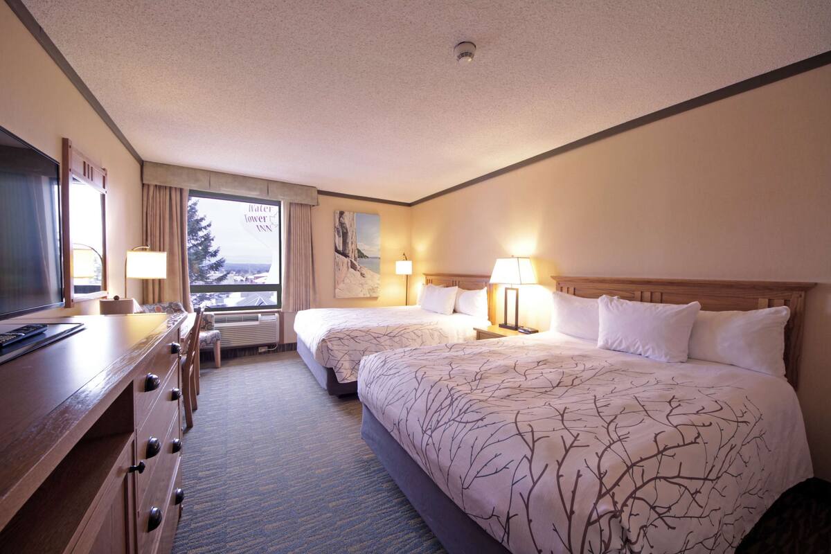 A hotel room with two beds, a desk with a TV, a window with a view of a snowy landscape, and a painting on the wall.
