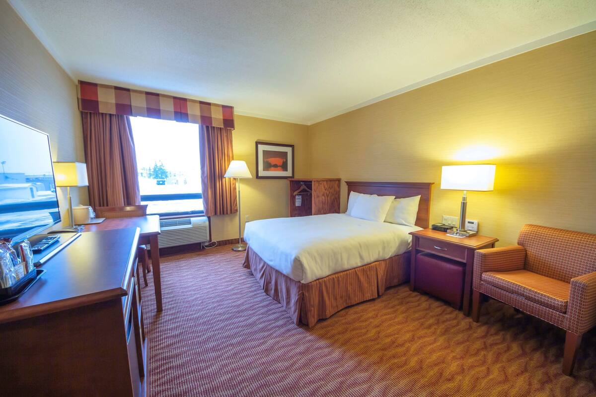 A hotel room with a bed, a desk, a chair, a nightstand, and a window with curtains.