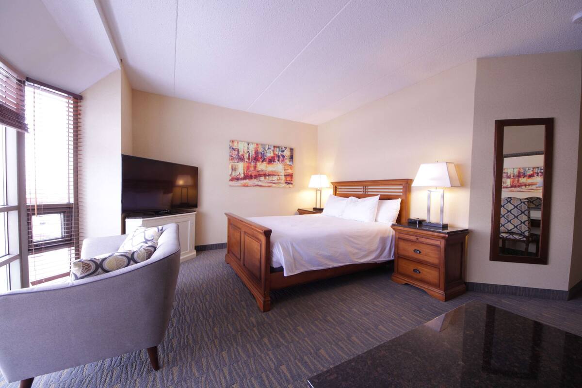 A hotel room with a bed, bedside tables, a lamp, a TV, a chair, and a painting on the wall.
