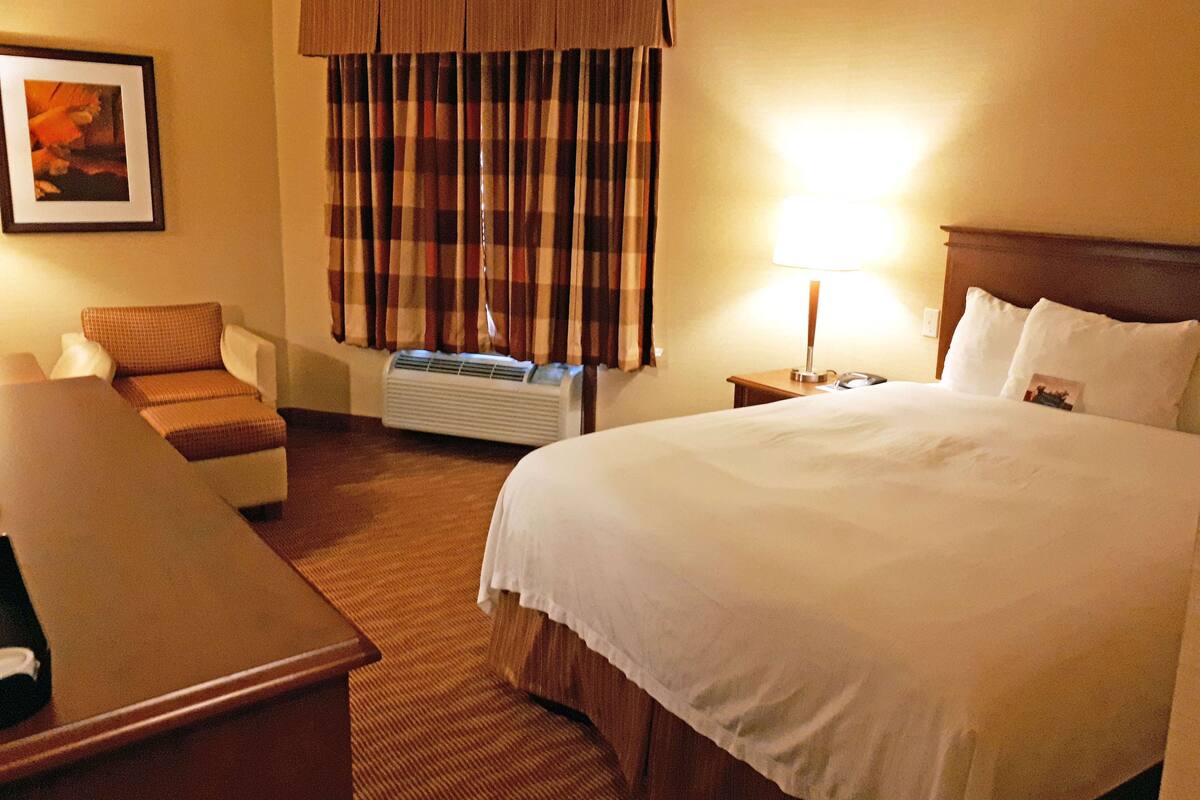 A hotel room with a large bed, a desk, a chair, a window with curtains, and a wall-mounted air conditioner.