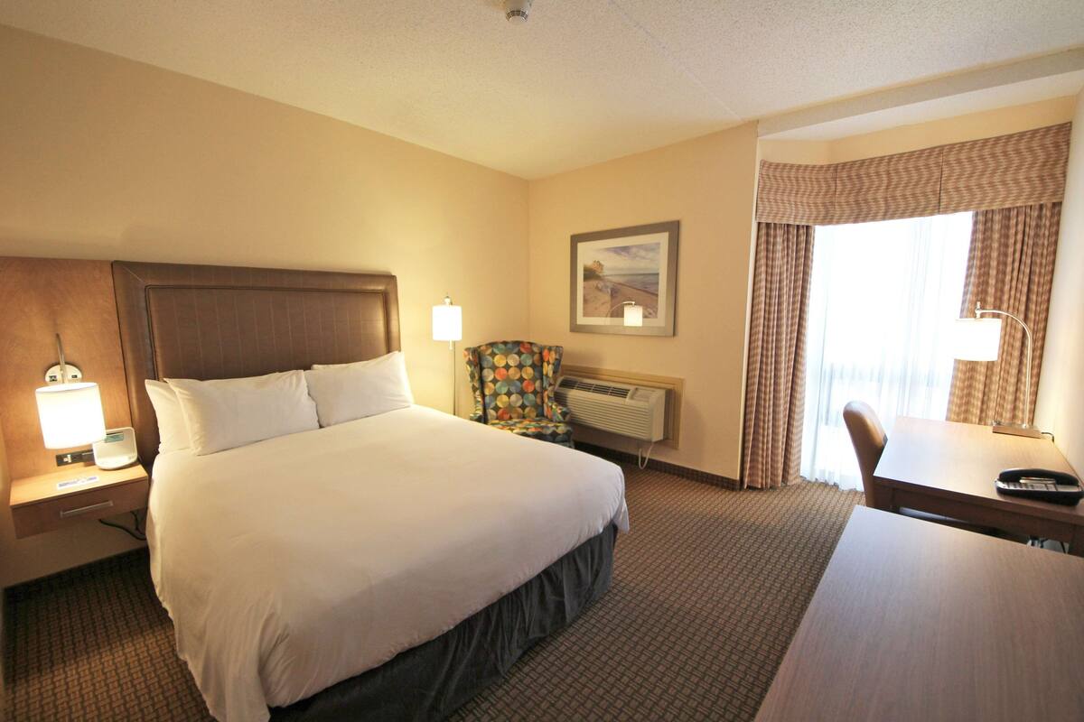 A hotel room with a large bed, a desk, a chair, a lamp, an air conditioner, and a window with curtains.