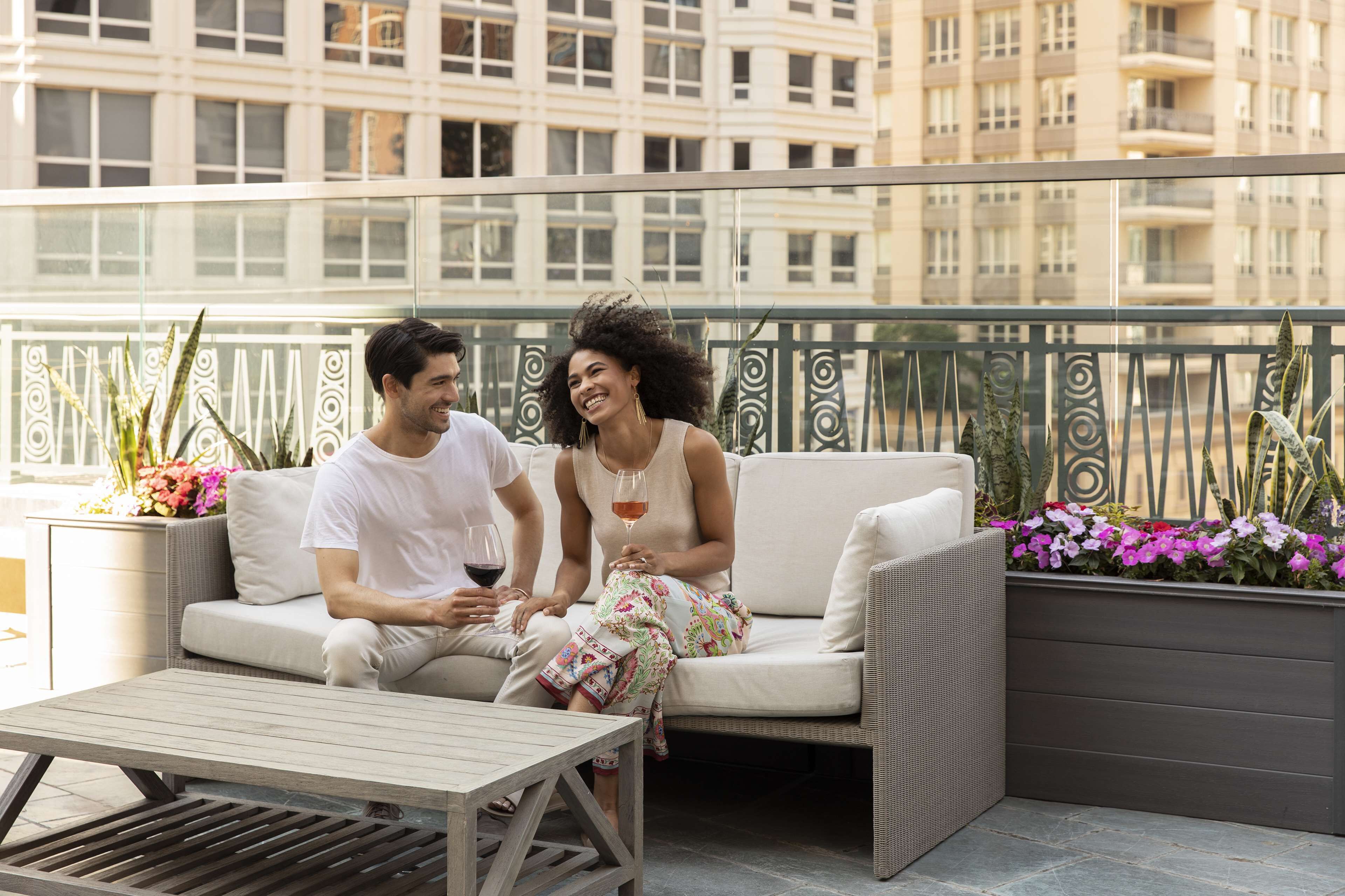 Un couple est assis sur un balcon d’où l’on a vue sur la ville. On y voit une table et des plantes en pot.