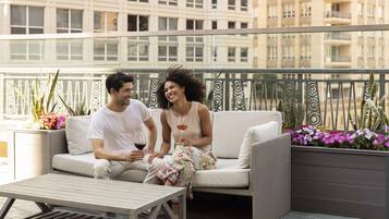 Un couple est assis sur un balcon d’où l’on a vue sur la ville. On y voit une table et des plantes en pot.