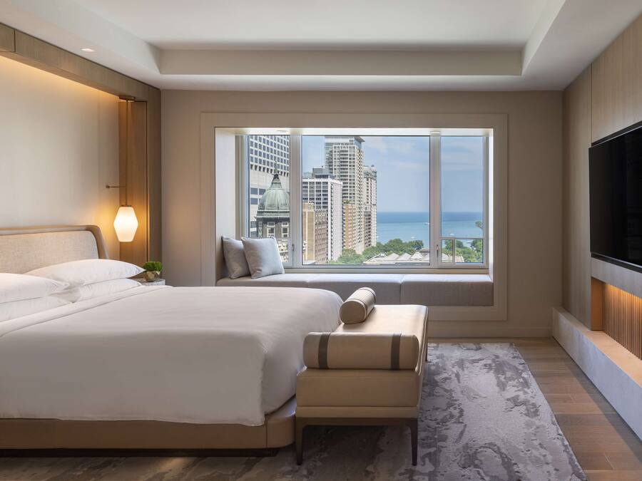 A modern hotel room with a large bed, a seating area, a TV, and a view of the city and ocean.