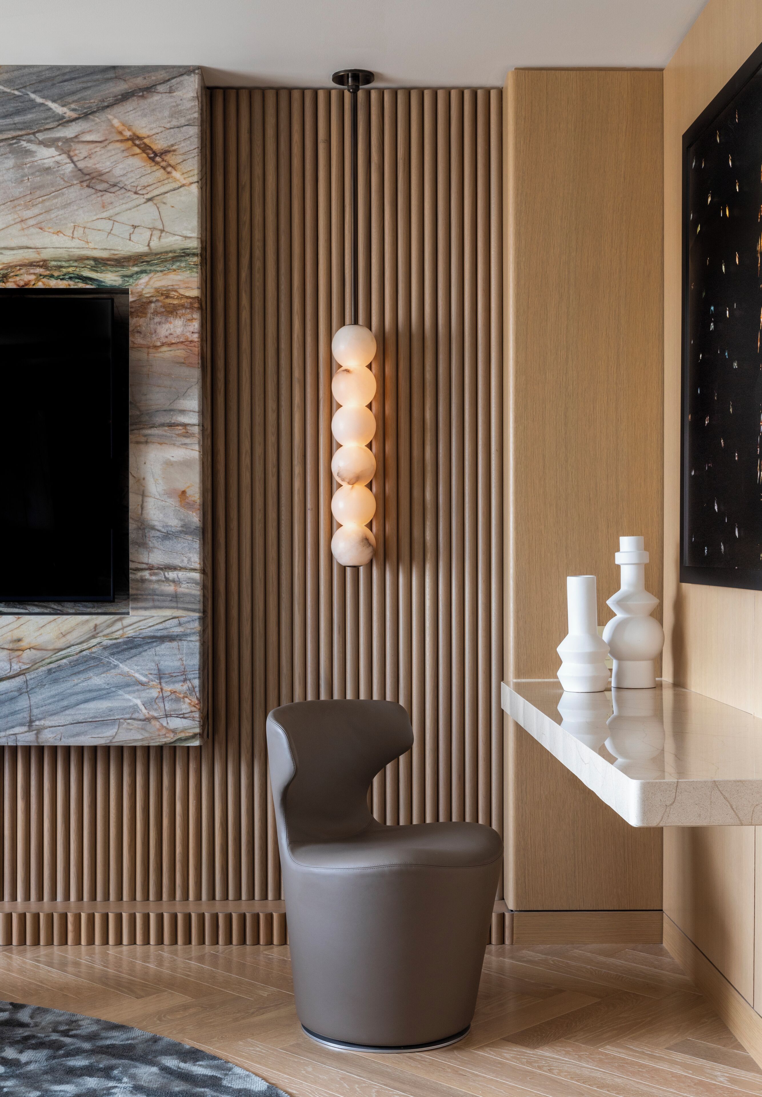 A modern interior with a flat-screen TV mounted on a marble wall, a wooden slatted partition, a round grey chair, and a white console table with decorative vases.