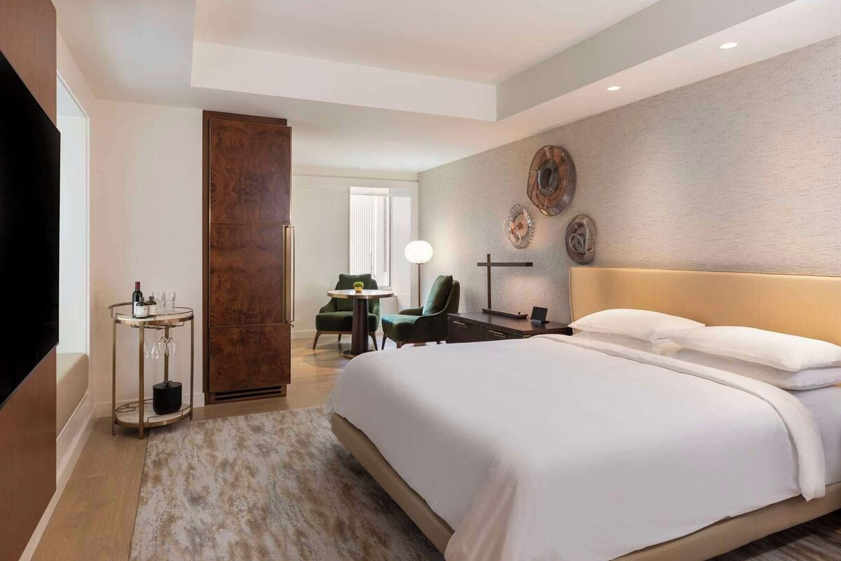 A modern hotel room with a large bed, a small round table with a bottle and glasses, a green armchair, a desk with a lamp, and decorative wall art.