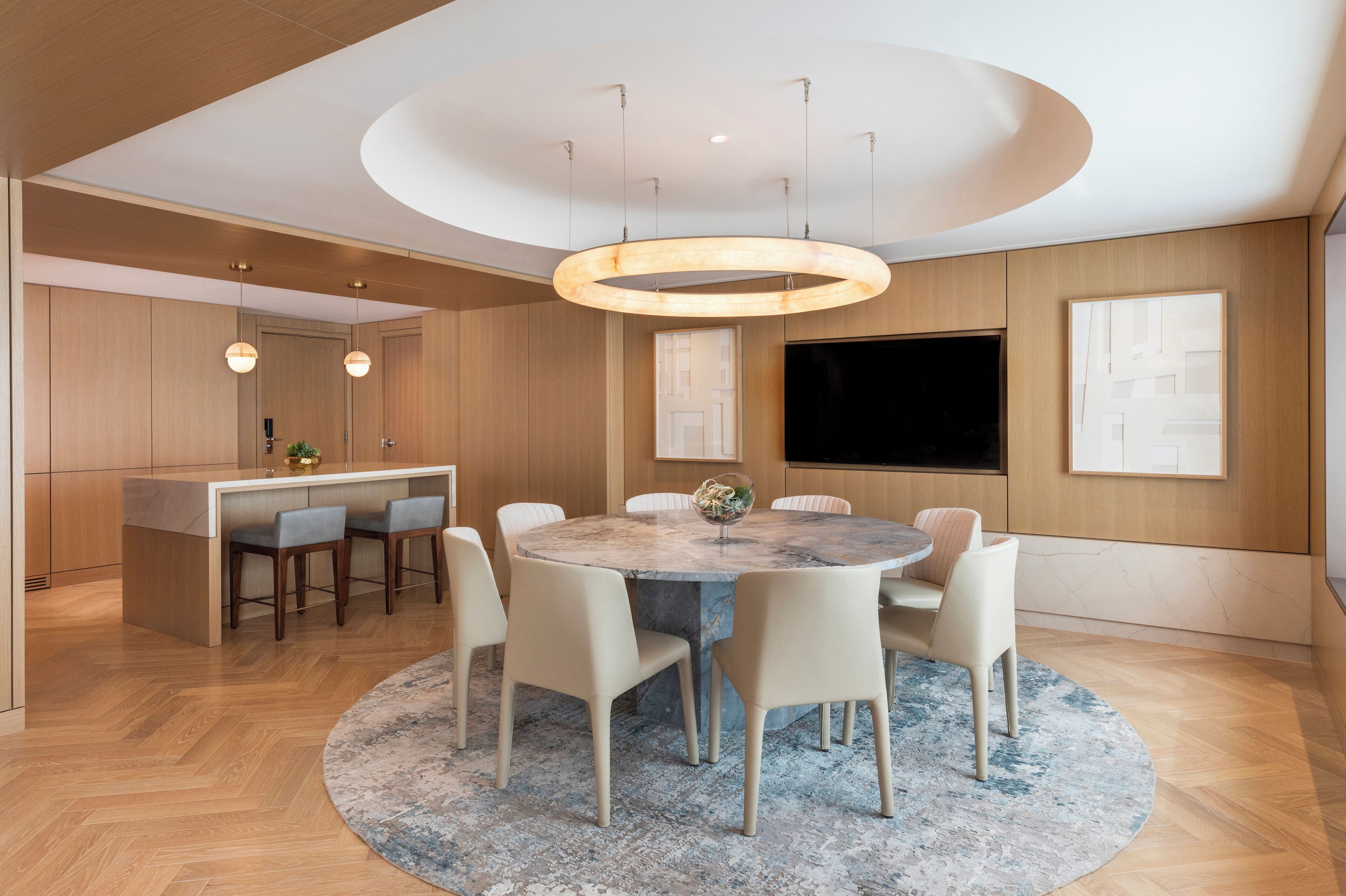 A modern kitchen with wooden cabinets, a central island with bar stools, a dining area with a marble table and chairs, and a large flat-screen TV mounted on the wall.