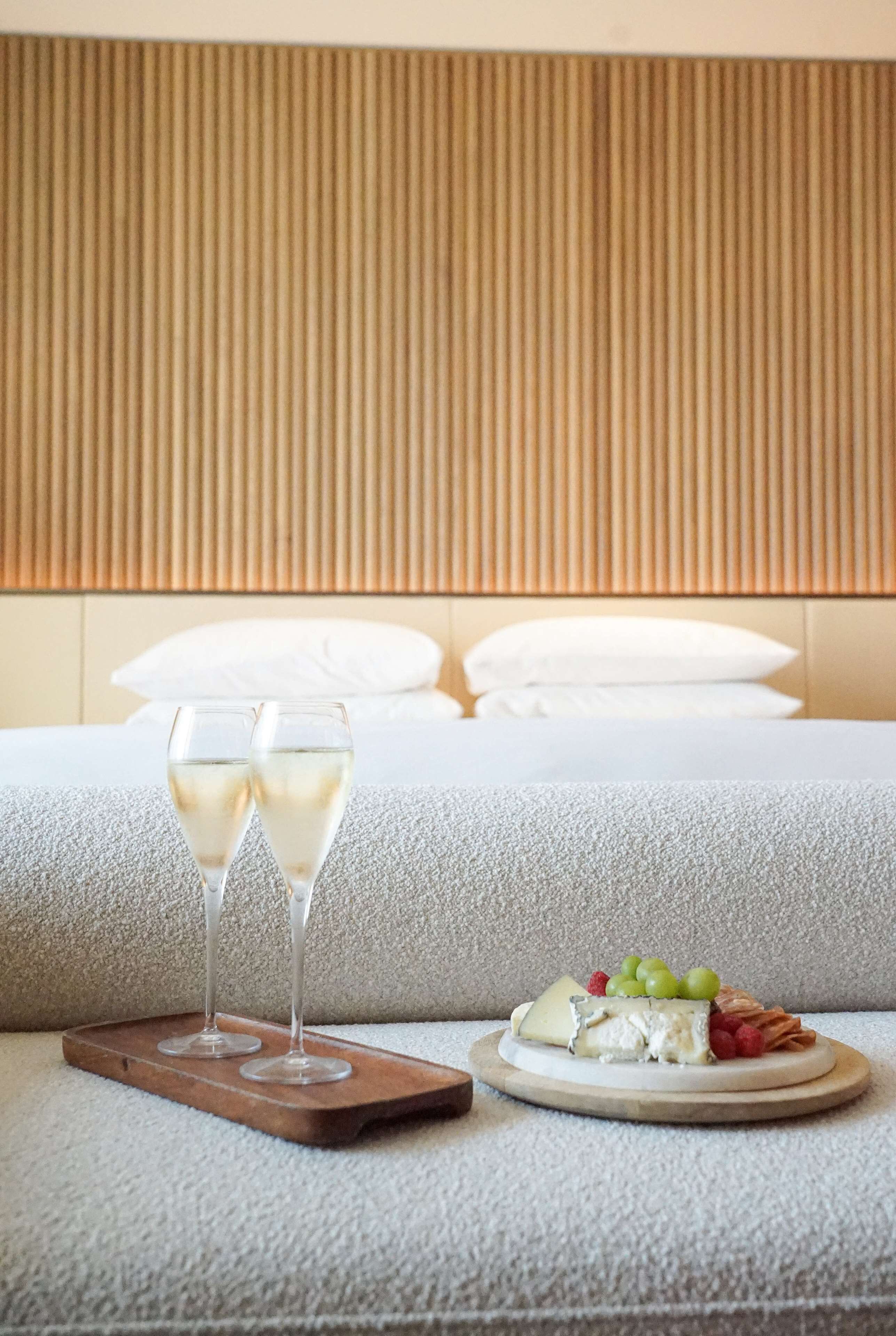 A bed with white linens, two glasses of white wine, and a cheese platter with grapes and crackers on a tray.