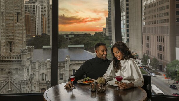 Um casal jantando à mesa com vista para a cidade e o pôr do sol.