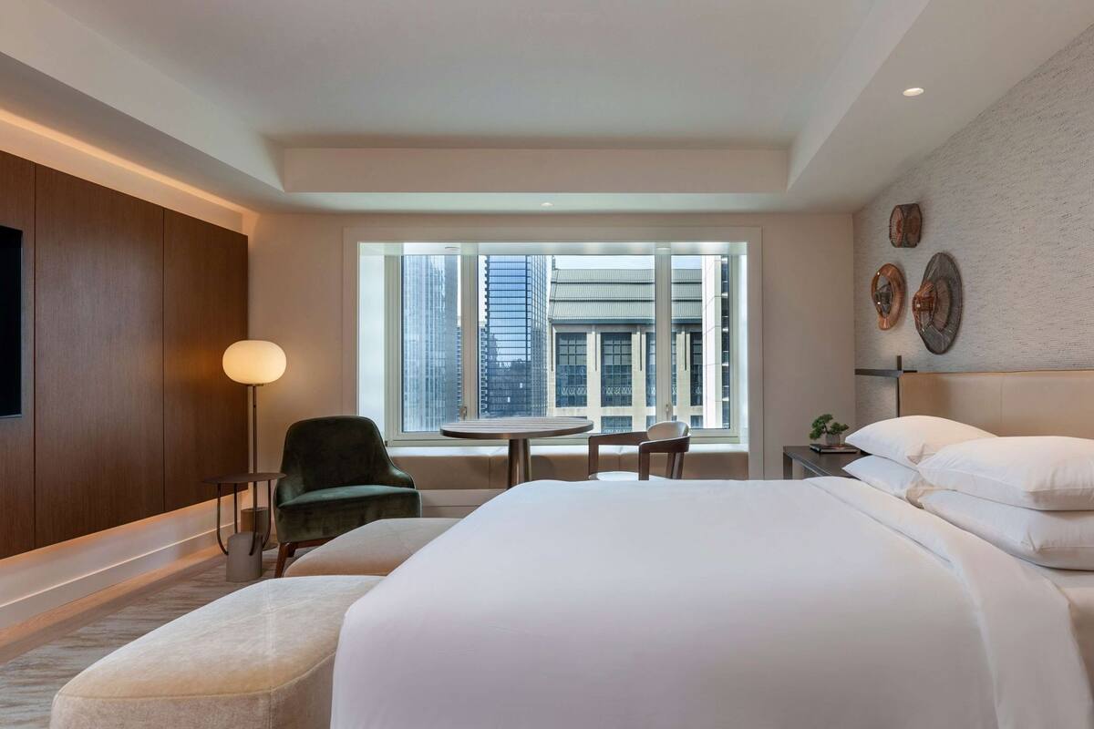 A hotel room with a large bed, a round table, two chairs, a lamp, and a view of tall buildings through a large window.