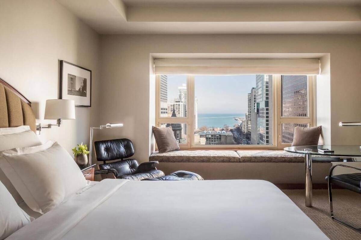 A hotel room with a large bed, a window seat with a view of the city, a desk, and a chair.