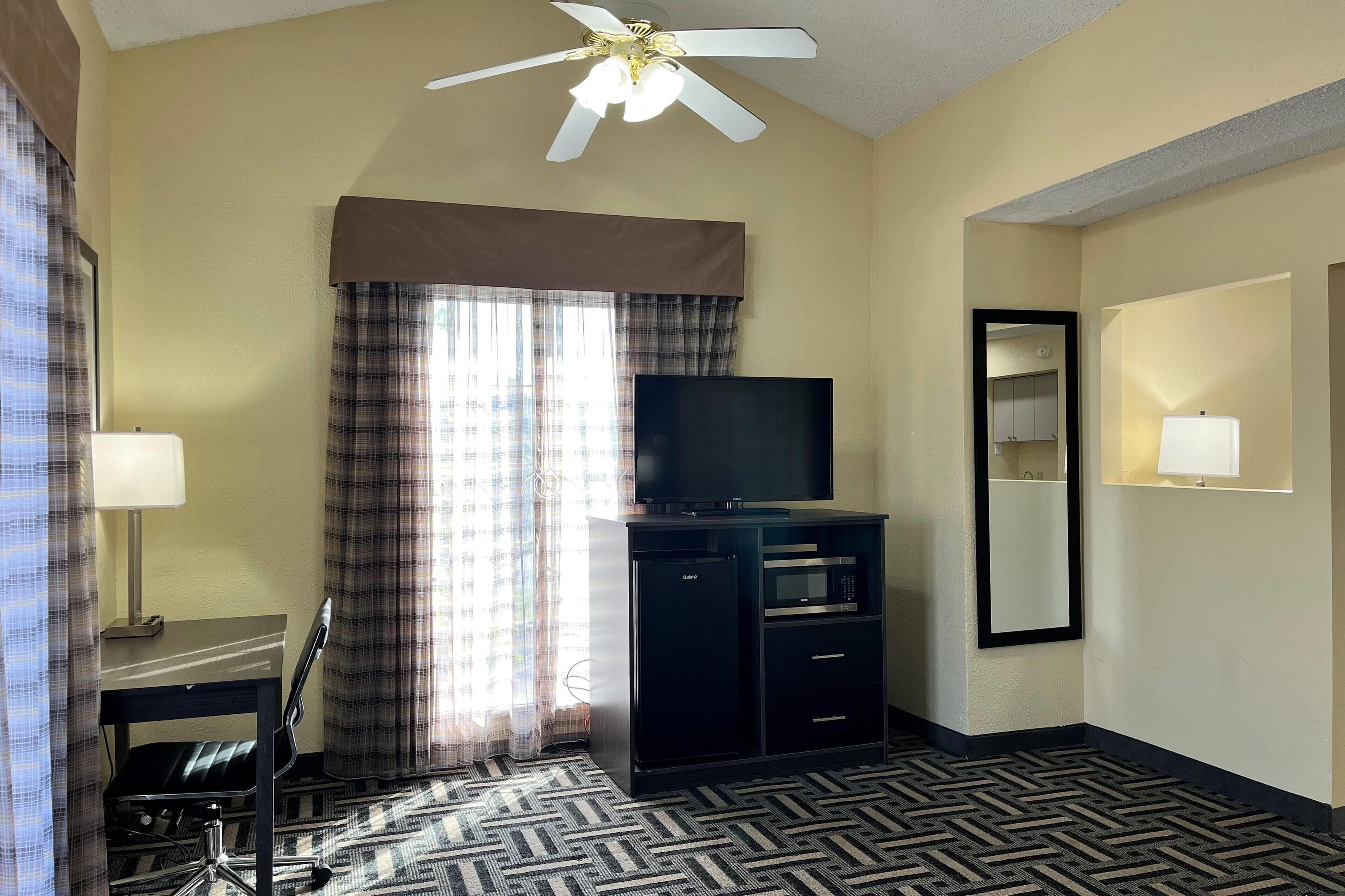 Une chambre d’hôtel avec un ventilateur de plafond, un téléviseur posé sur un meuble, un bureau avec une chaise, une moquette à motifs et une fenêtre avec des rideaux.