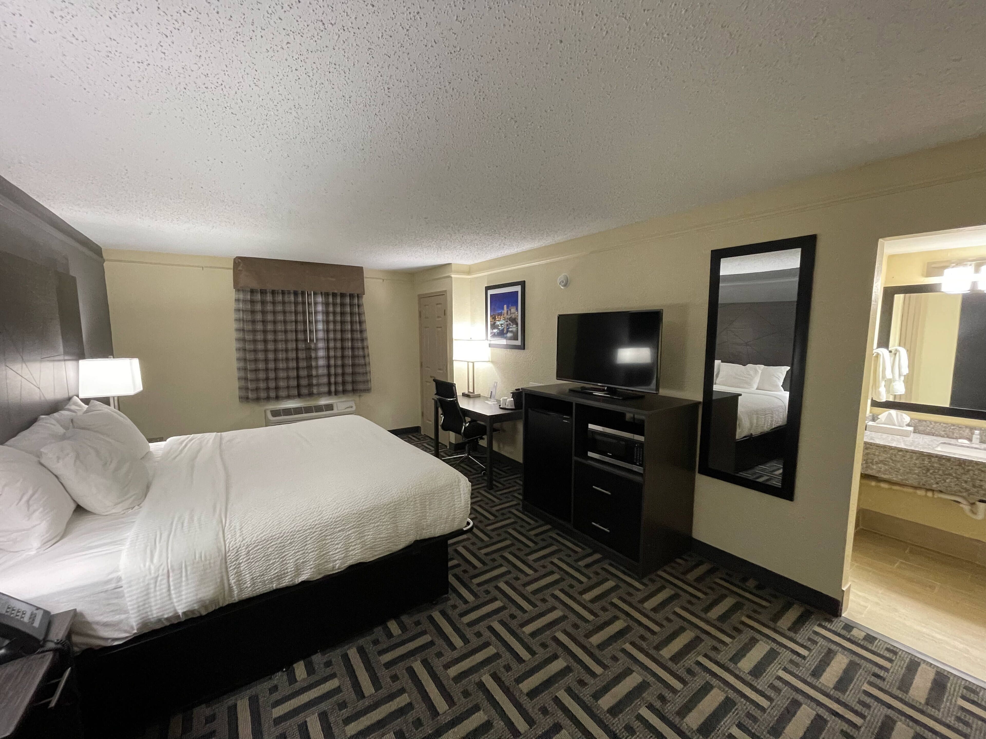 A hotel room with a bed, a desk with a television, a mirror, and a bathroom with a sink and mirror.
