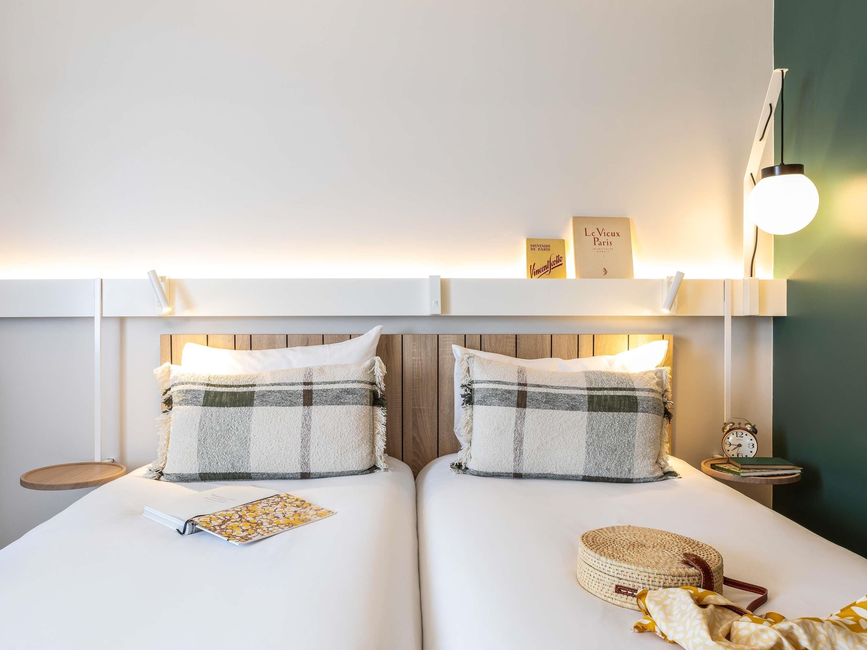 Una cama doble con dos almohadas, una mesita de noche con un libro y una lámpara, y una estantería de pared con libros.