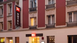 Eine Hotelfassade mit einem gut sichtbaren 'ibis HOTEL'-Schild, mehreren Balkonen und einem Schild mit der Aufschrift 'Maine Montparnasse', das den Standort angibt.