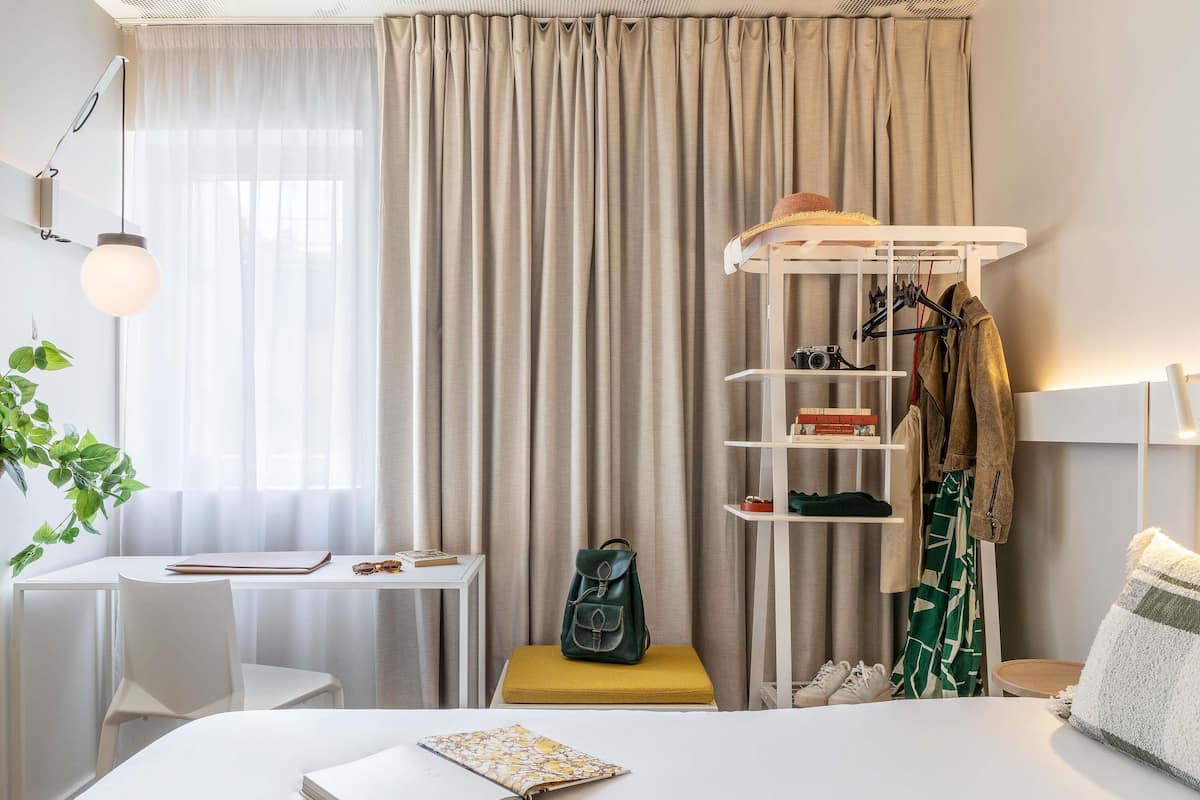 Una habitación de hotel ordenada con cama, escritorio, silla, zapatero colgante y planta en maceta.