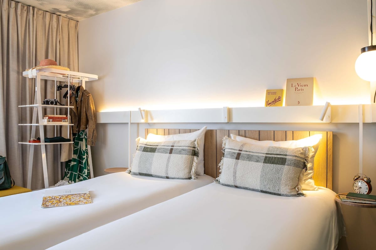 Una habitación de hotel con una cama, una estantería con ropa y una cámara, un reloj y un cartel en la pared.