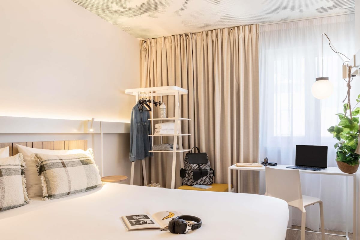 Una habitación de hotel ordenada con una cama, una mesita de noche, un escritorio con una computadora portátil, una planta colgante y un armario.