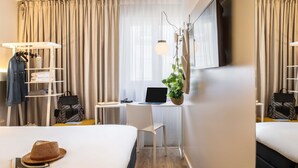 Ein Hotelzimmer mit einem Bett, einem Schreibtisch mit Laptop, einer hängenden Pflanze und einem Kleiderschrank mit Kleidung.