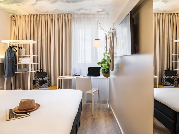 A hotel room with a bed, a desk with a laptop, a hanging plant, and a wardrobe with hanging clothes. - ibis Paris Maine Montparnasse 14ème (Paris)