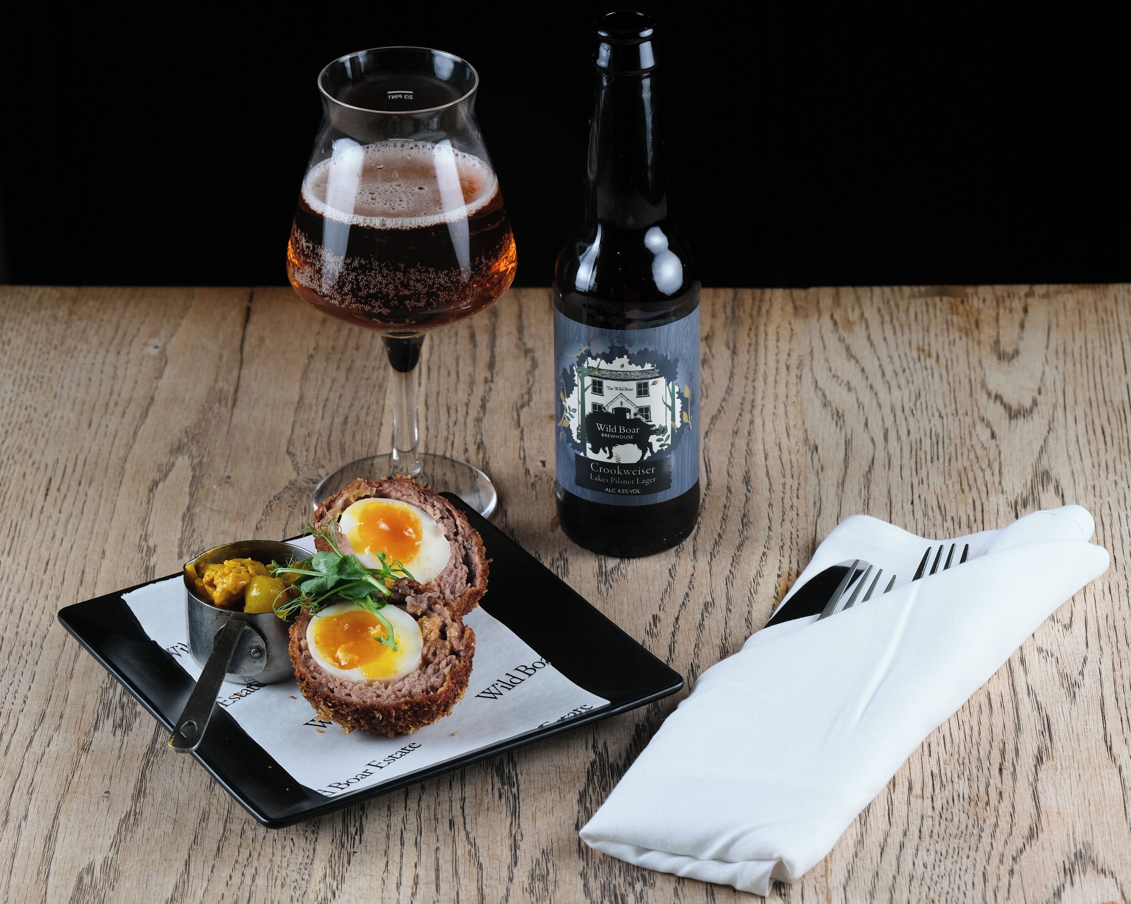 A plate with a Scotch egg, a glass of beer, and a bottle of beer on a wooden table.