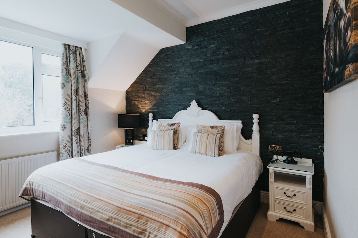 A bedroom with a large bed, a dark accent wall, a window with curtains, and a nightstand with a lamp.